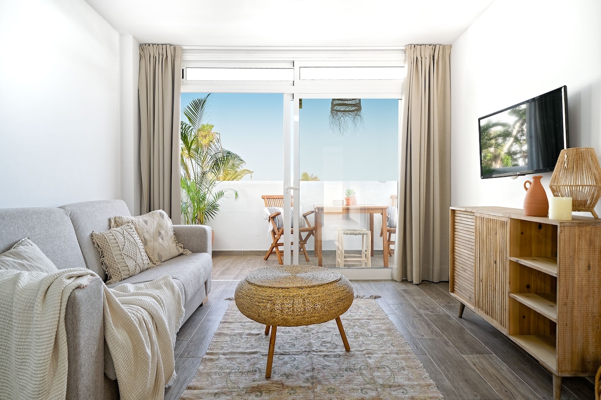 A bright living area features a comfortable sofa with decorative cushions and a circular coffee table. Large glass doors provide a view of the outdoor seating area, framed by lush greenery. Natural light enhances the neutral color palette and warm wooden accents throughout the space.