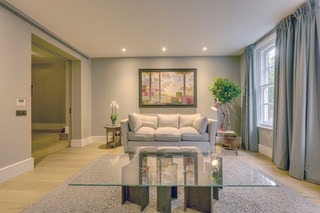 A well-lit living area features a grey sofa positioned against a neutral wall, complemented by a decorative painting above. A glass coffee table occupies the center, and a potted plant adds a touch of greenery. Light curtains frame the window, contributing to the room’s brightness.