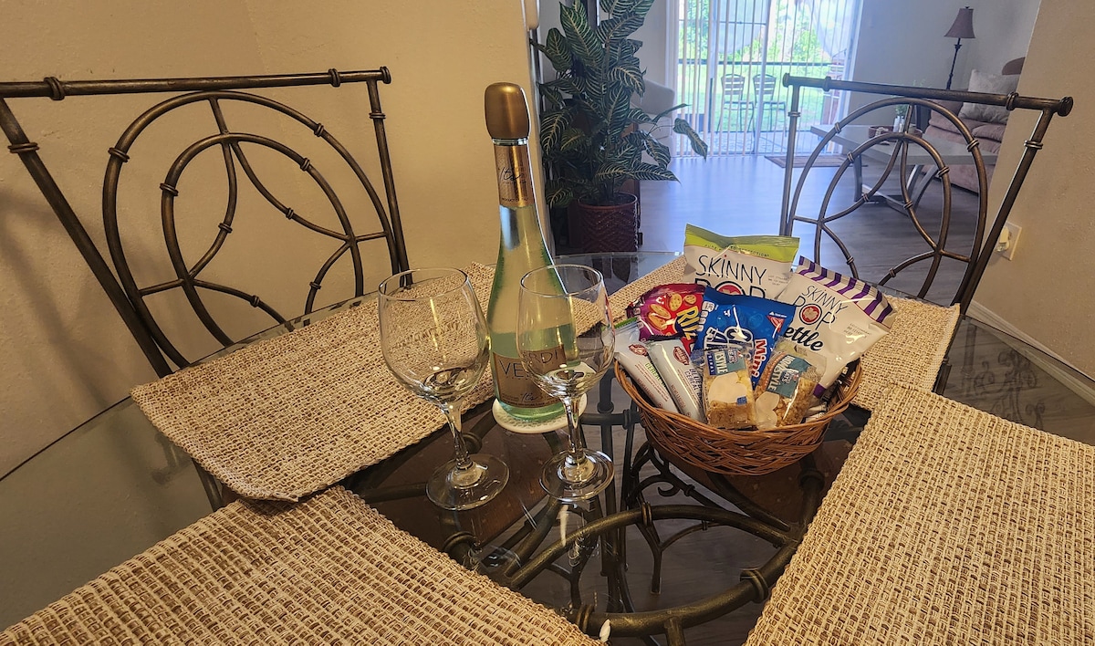 A dining table is set with a glass bottle of sparkling wine and two elegant glasses. A woven basket contains assorted snacks, surrounded by placemats. In the background, a glass door opens to a sunny outdoor space, framed by greenery.