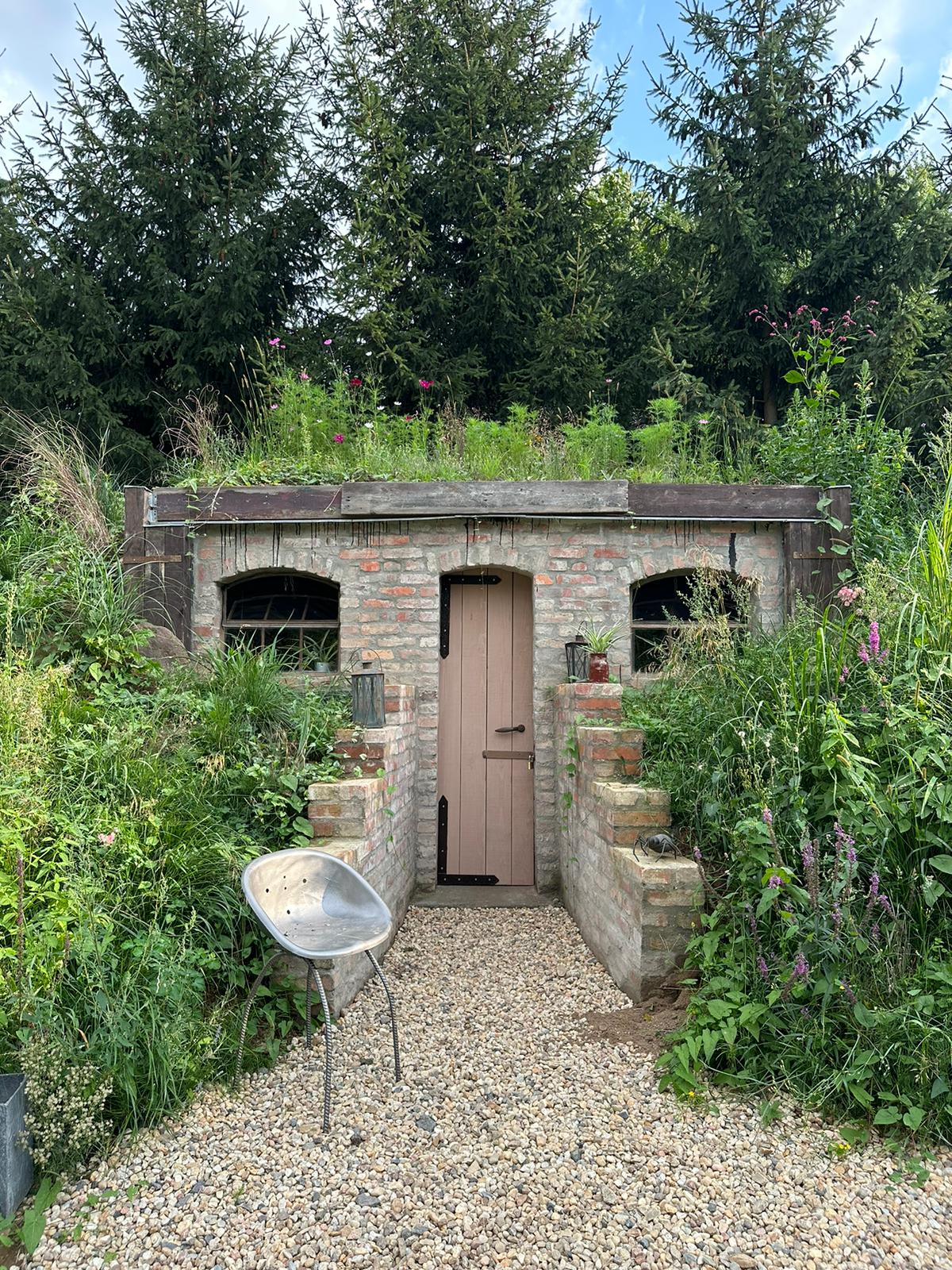 The entrance of a rustic earth house is framed by vibrant greenery and wildflowers. A path of gravel leads to a modest wooden door, flanked by lamps and steps that provide access from the surrounding garden.