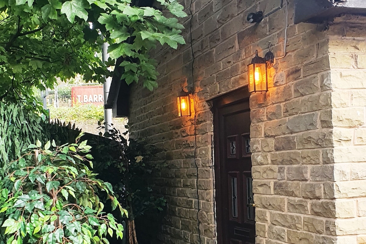 The entrance to the Coach House is framed by lush greenery, with warm lights illuminating the stone exterior. A solid wooden door is set into the wall, providing a welcoming access point to the property.