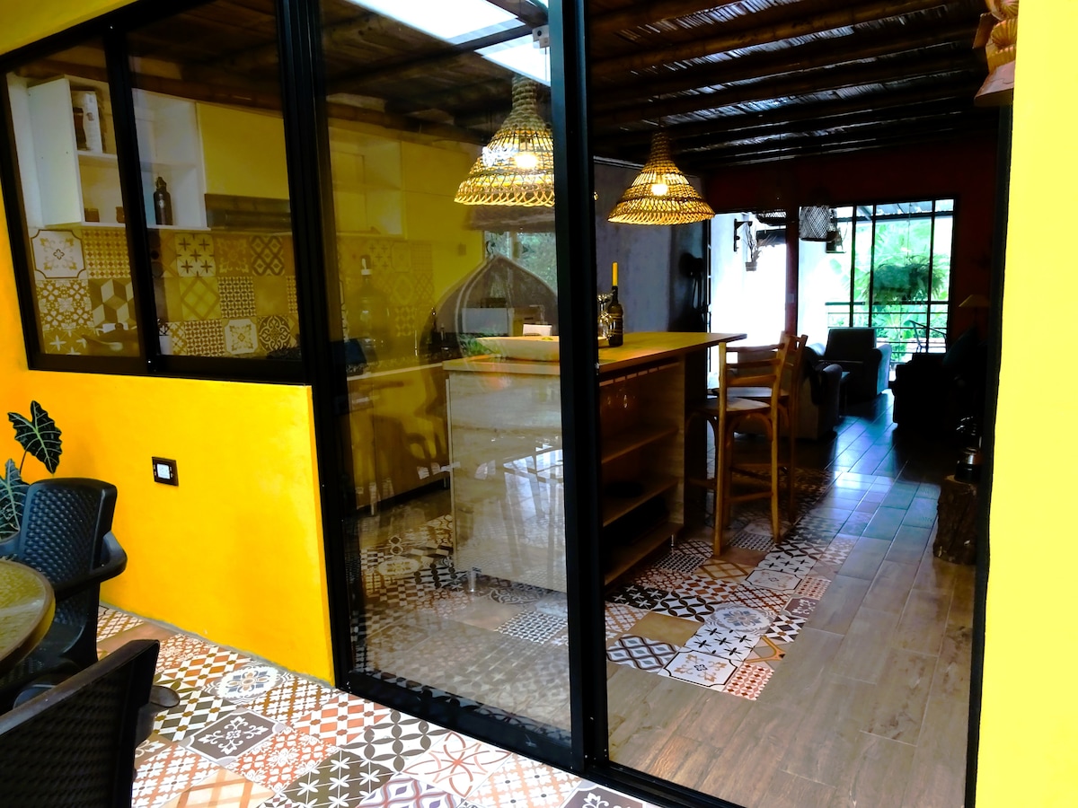 An open kitchen area showcases colorful tile flooring with a variety of patterns. Natural light floods the space through large glass doors. Rattan pendant lights hang above a wooden island, while shelves display decor items in a vibrant atmosphere.