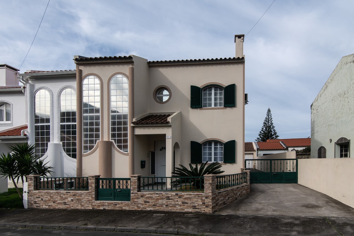 The exterior of the house features a charming facade with arched windows and green shutters. A low stone wall encloses the front yard, which is complemented by small plants and a paved driveway leading to the gated entrance.