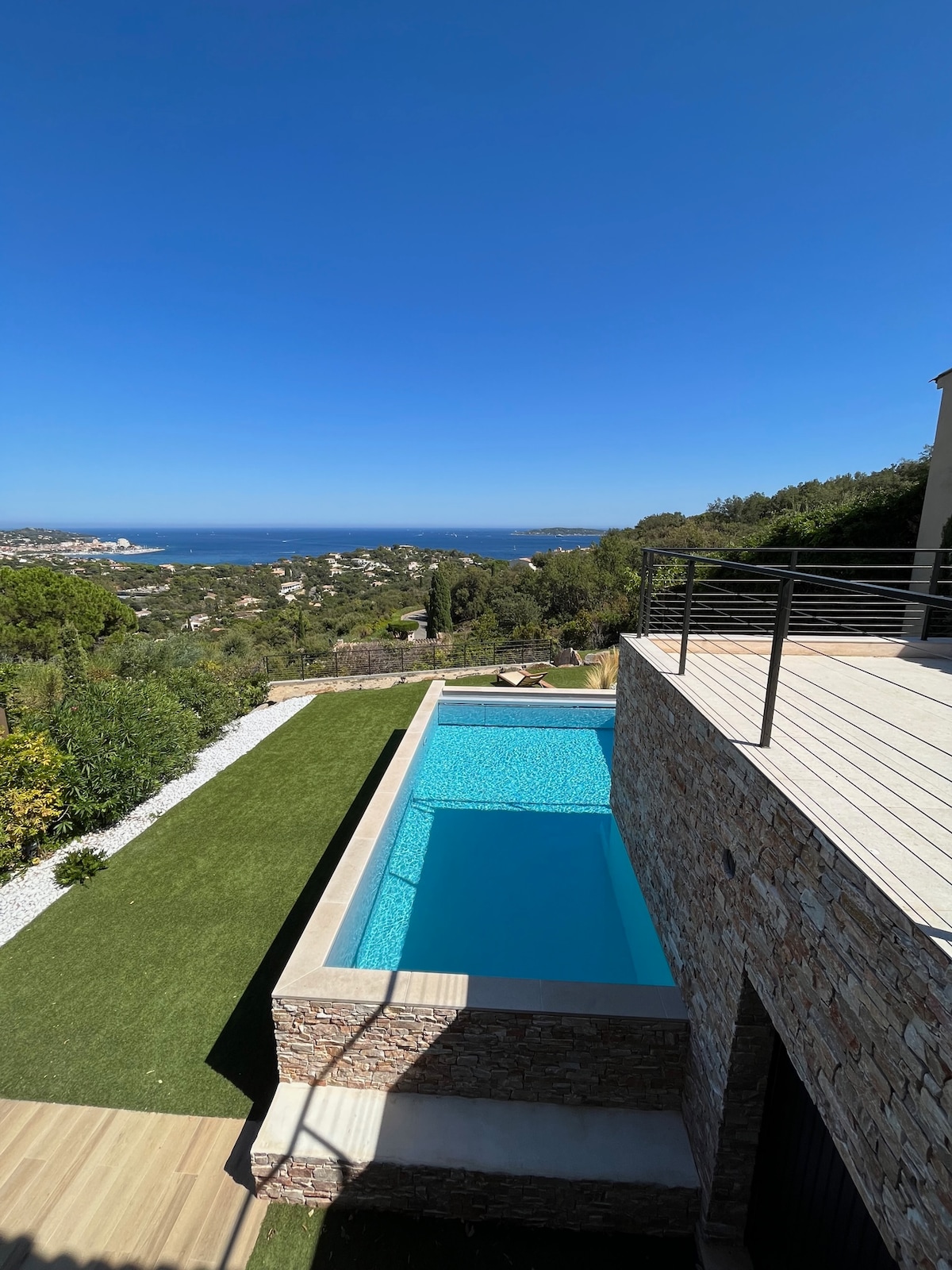 A modern pool is set on a stone terrace, surrounded by manicured greenery and offering views of the expansive landscape and the ocean beyond. The bright blue water of the pool contrasts with the clear sky, creating a serene outdoor environment.