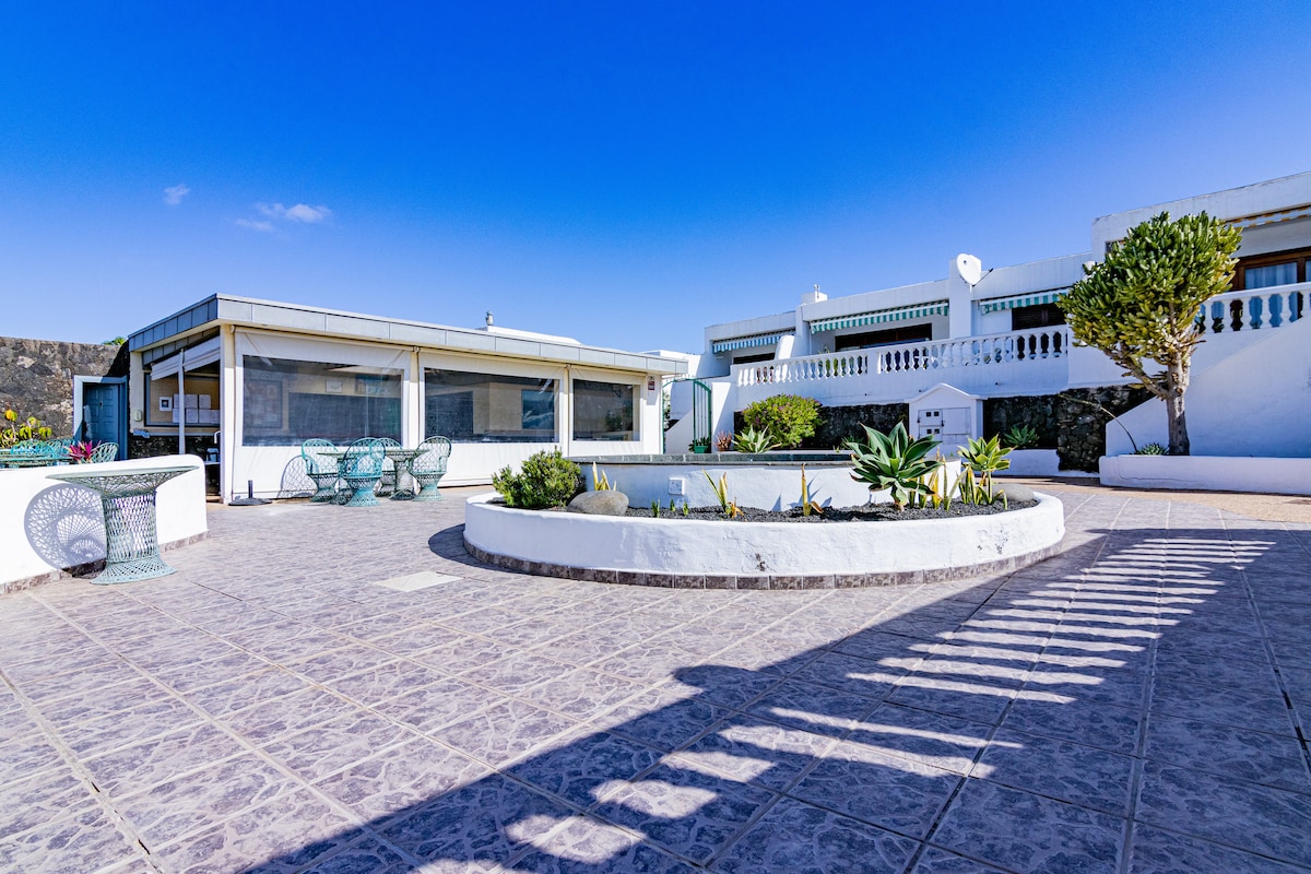 A spacious outdoor area is presented, featuring a circular garden bed with succulents and a stone pathway. Comfortable seating arrangements made from wicker are visible, surrounded by a clean, tiled surface beneath a clear blue sky.