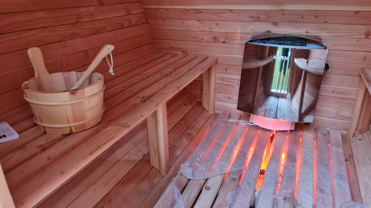 The sauna interior features wooden benches along the walls, offering a natural and warm atmosphere. A metal heater is positioned on one wall, with reflective surfaces complementing the wood paneling. A wooden bucket, along with a ladle, is placed nearby, creating a calming spa-like environment.