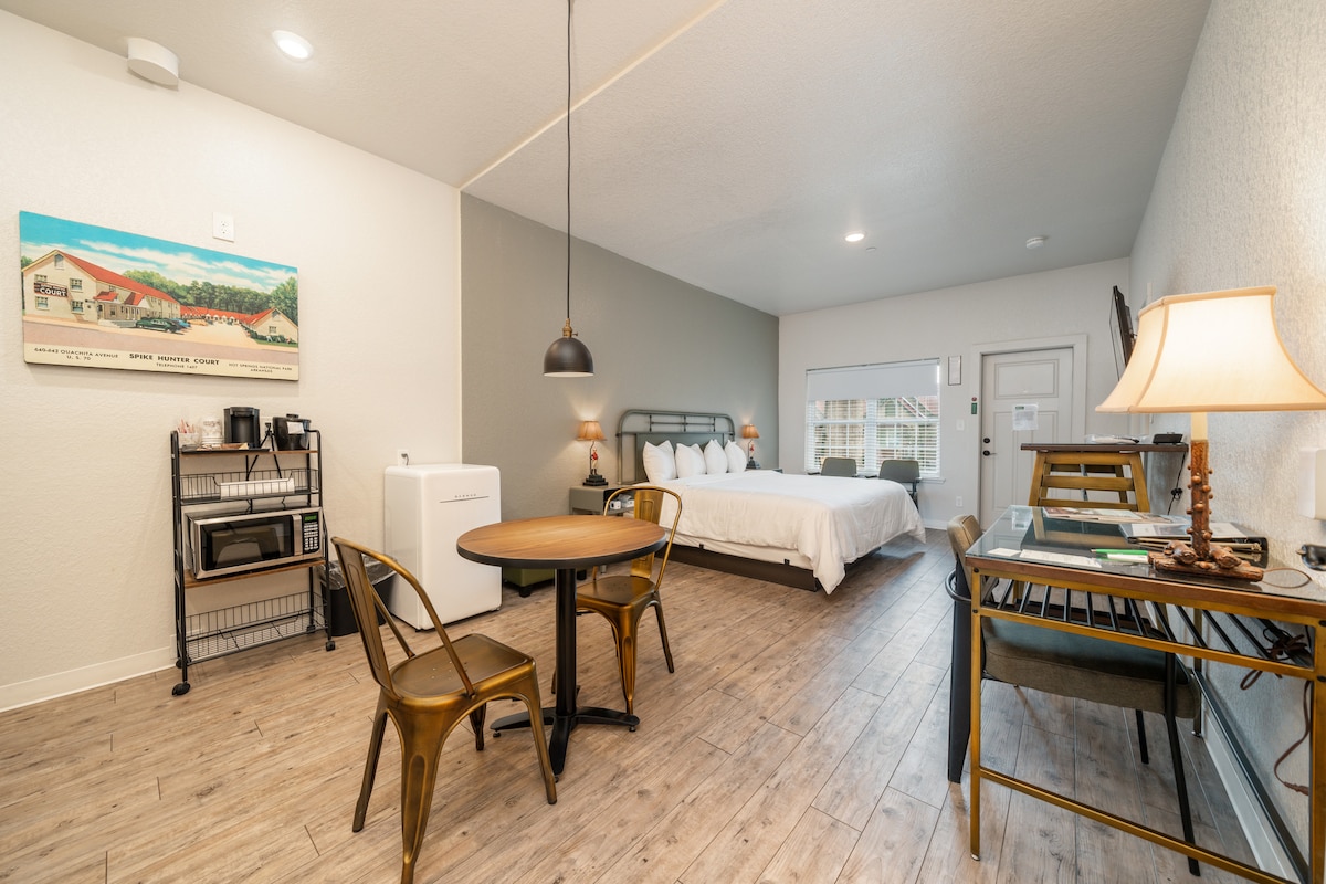 A spacious studio suite is displayed, featuring a comfortable king bed with white linens. A wooden dining table is paired with four metal chairs. A kitchenette area is visible with a mini refrigerator, microwave, and coffee maker. Natural light enters through the window, enhancing the inviting atmosphere.