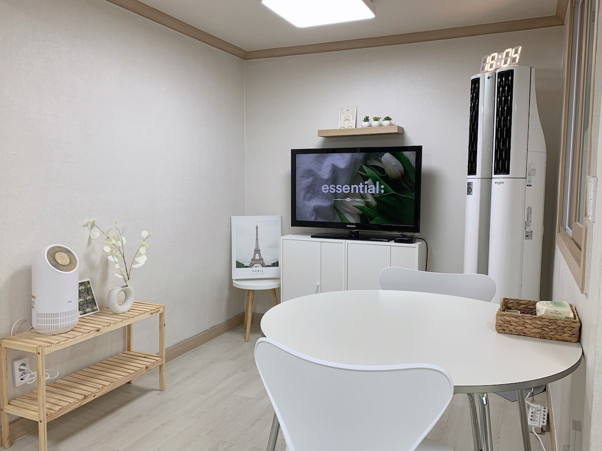 A simple living area features a light-colored wall and flooring. A white table with chairs occupies the center, accompanied by a shelf with a decorative vase. A television displays a minimalistic screen, while an air conditioning unit stands beside a white cabinet.
