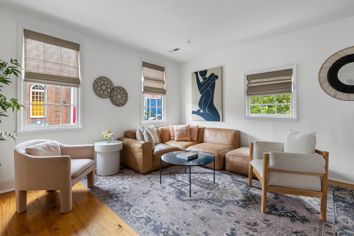 A modern living area is presented with a curved chair and a comfortable leather sectional. Light filters in through windows adorned with woven shades. Decorative wall art and a round mirror add character, while an area rug defines the space, contrasting with the warm wood flooring.