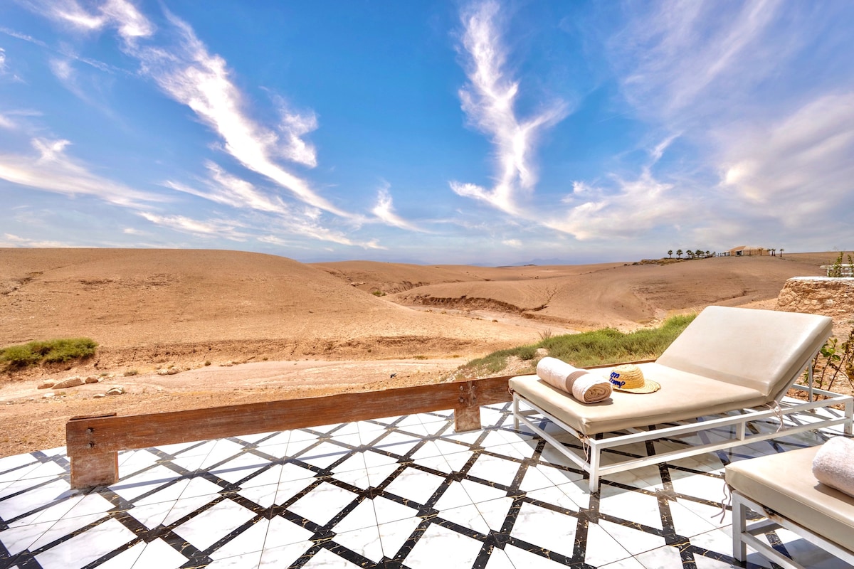 A sunlit terrace features two lounge chairs positioned on a patterned tile floor. The view overlooks the expansive desert landscape, with rolling dunes and a clear blue sky adorned with wispy clouds.