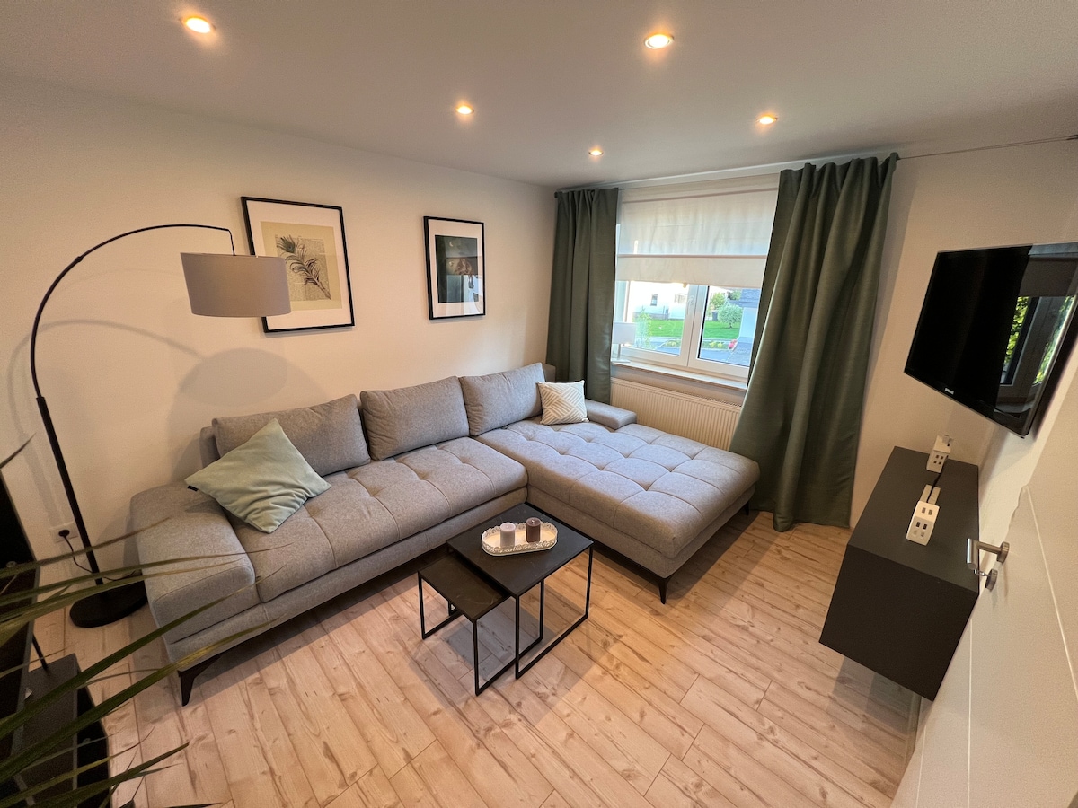 A spacious living area features a modern gray sectional sofa with a light teal accent pillow. A compact black coffee table sits in front of the sofa, accompanied by decorative items. Natural light enters through a large window, complemented by green curtains, creating a bright atmosphere.