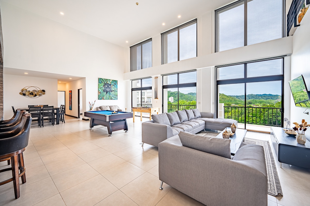 A spacious living area features large windows that fill the room with natural light, showcasing views of the surrounding mountains. Comfortable seating is arranged around a sleek coffee table, and a dining area with a modern table is seen in the background. The room offers a stylish and open design.