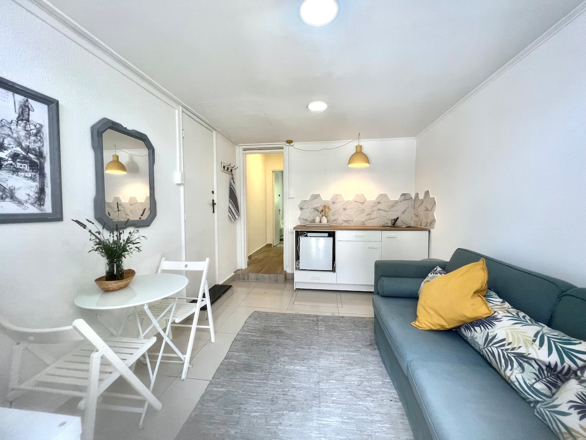 The living space features a minimalist design with a blue sofa, complemented by a yellow pillow. A small dining area with two white chairs is set beside a kitchenette. Natural light enters through the windows, enhancing the bright, neutral colors of the decor.