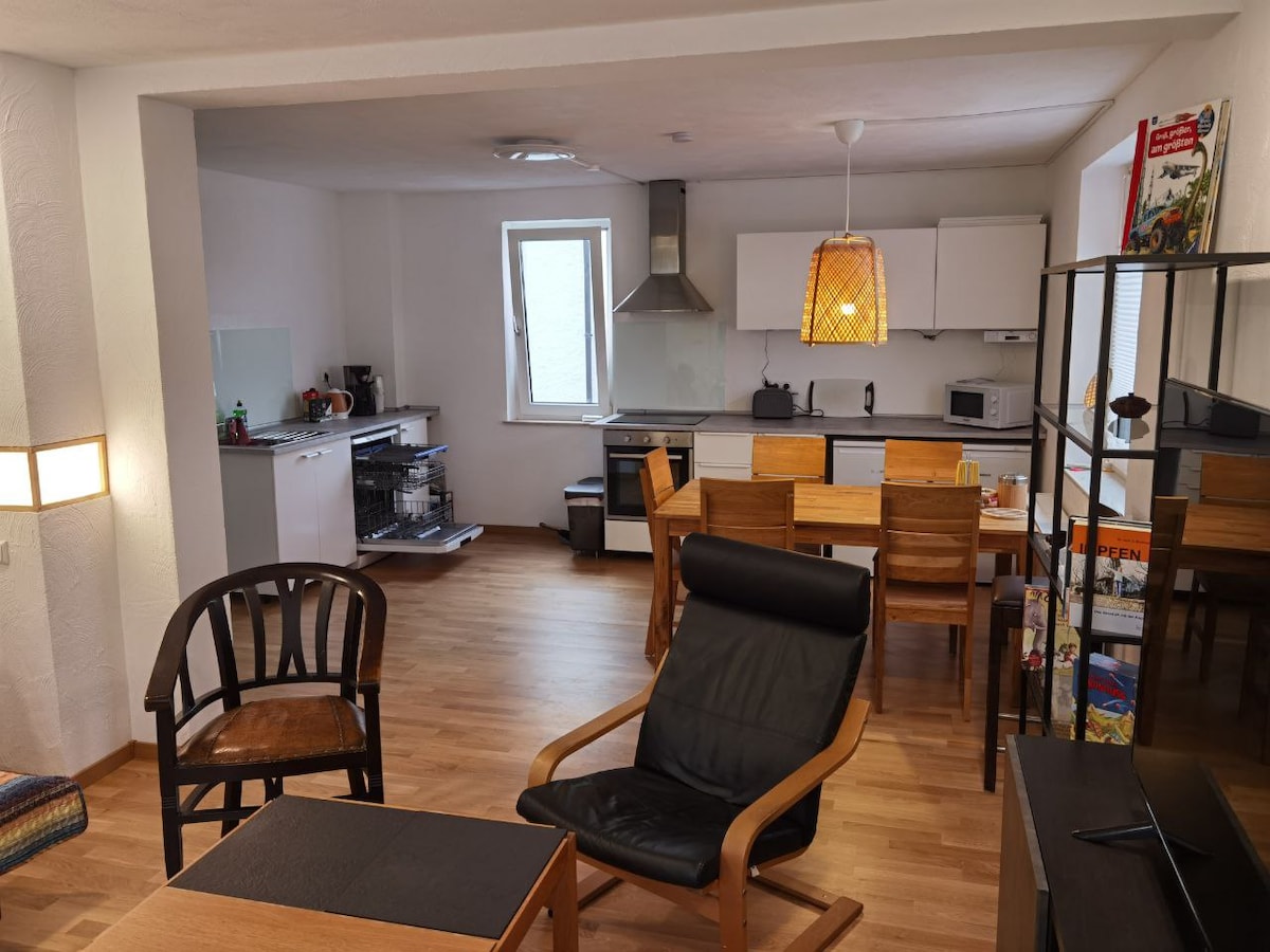 An open kitchen and dining area features modern appliances, a dining table with wooden chairs, and a cozy seating arrangement. Natural light enters through a nearby window, enhancing the warm wooden flooring and neutral wall colors.