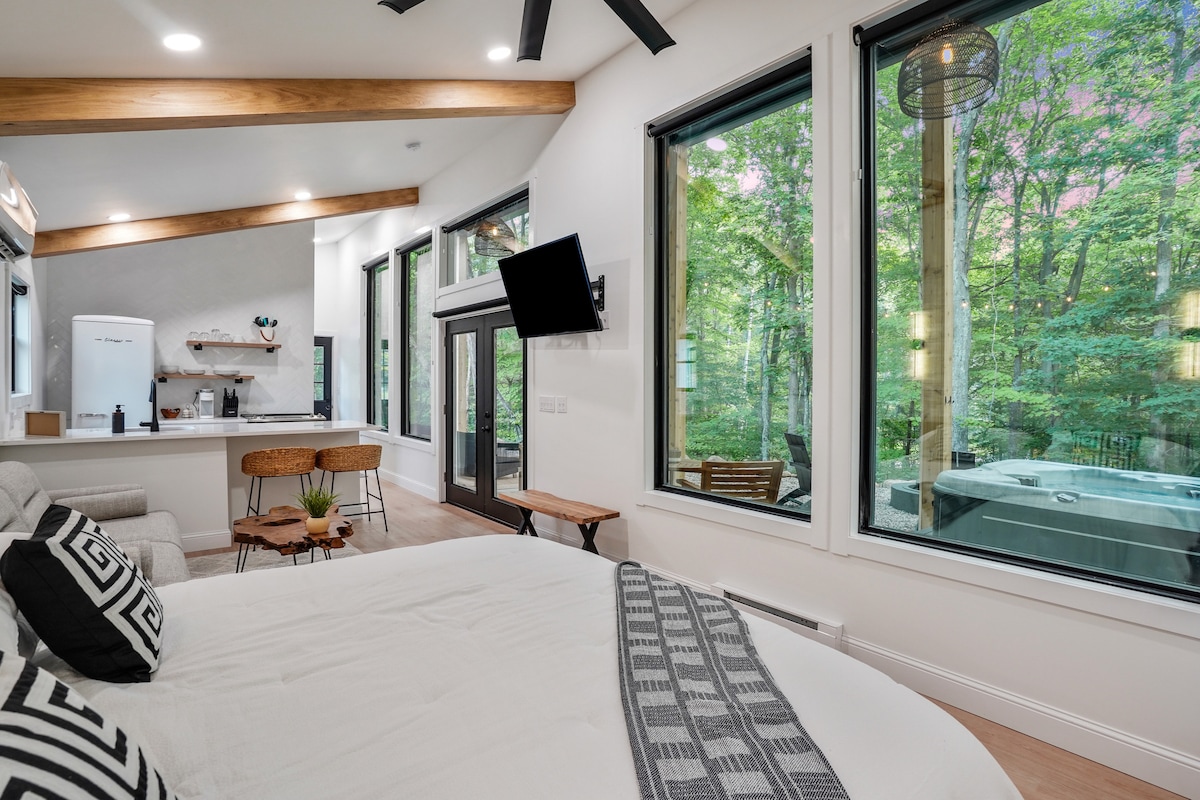 A spacious living area is featured with large floor-to-ceiling windows that provide views of the surrounding woods. A comfortable king-size bed is positioned prominently, accompanied by modern furnishings and a cozy throw. A glimpse of the private hot tub is visible through the glass doors.