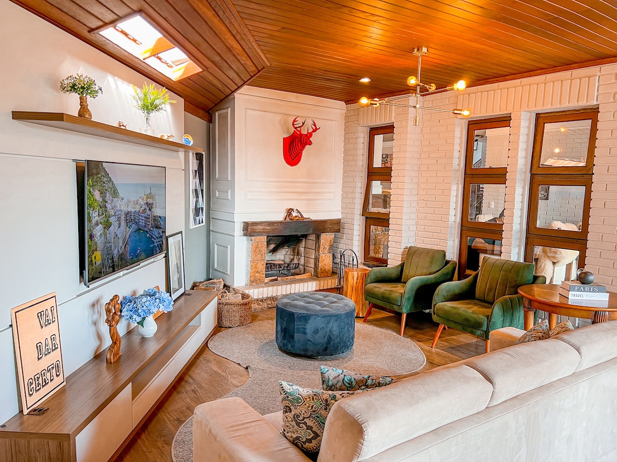 A cozy living area features a large cream sofa alongside two green armchairs, creating a relaxed seating arrangement. A round ottoman sits in the center on a soft rug. A wooden entertainment unit houses a television, while a fireplace adds warmth to the space.