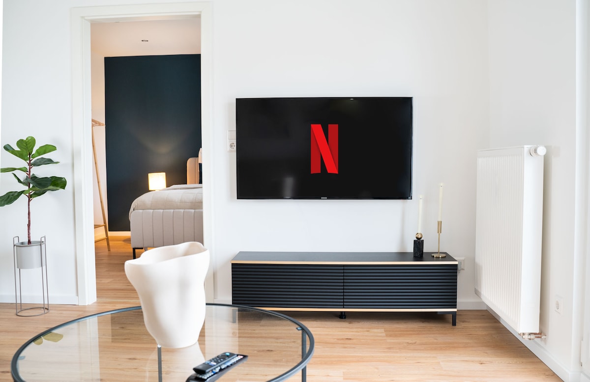 A bright living area features a sleek black media console with a wall-mounted Smart TV displaying the Netflix logo. A glass coffee table is positioned in the foreground, while a plant adds a touch of greenery. The doorway leads to a softly lit bedroom.