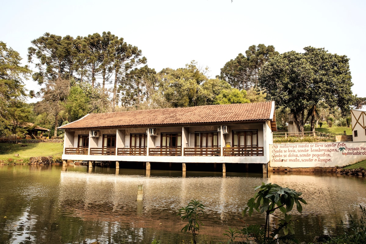 A serene building is set on stilts by a calm lake, featuring a traditional tiled roof and multiple windows. Lush greenery surrounds the structure, creating a tranquil environment. Reflections of the building and trees can be seen in the still water.