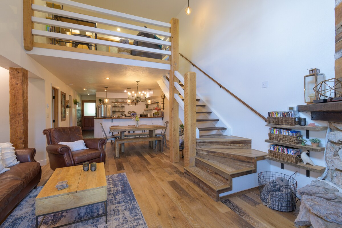 An inviting living area displays warm wooden accents with a comfortable leather sofa and a rustic coffee table. A staircase leads to an upper loft area, with a dining space visible in the background. Features include a chandelier and open shelving along the walls.