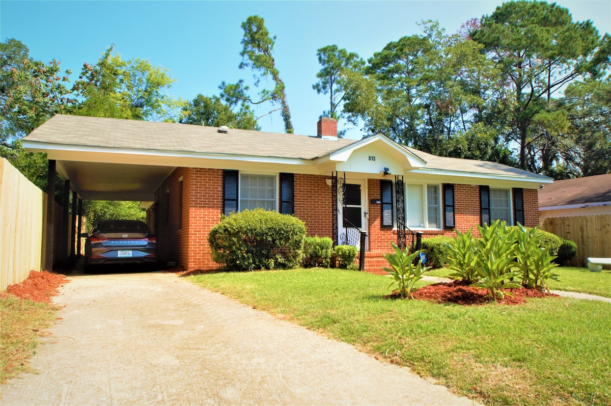 A single-story brick home is presented with a neat front yard featuring lush greenery and well-maintained shrubs. A carport accommodates vehicles beside a broad driveway. The entrance is framed by a simple awning and flanked by window shutters, creating an inviting atmosphere.