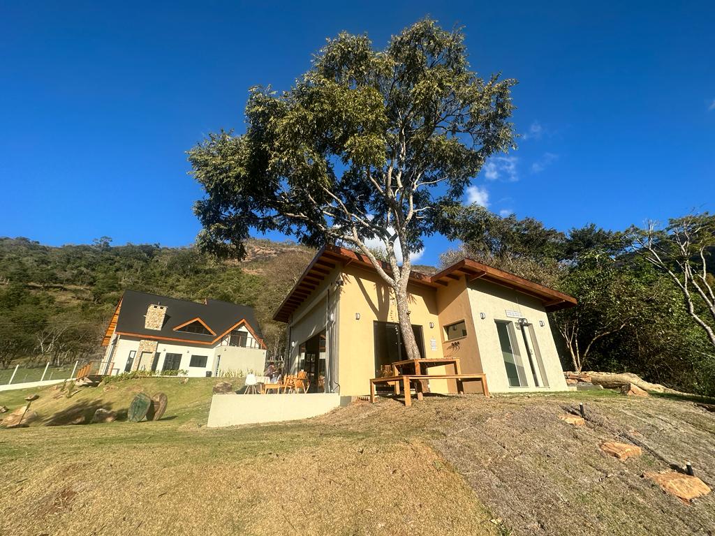A rustic and newly constructed chalet is showcased, surrounded by greenery and hills. The property includes large windows and a welcoming outdoor dining area, offering space for relaxation. A tree provides shade while the blue sky creates a serene backdrop for the setting.