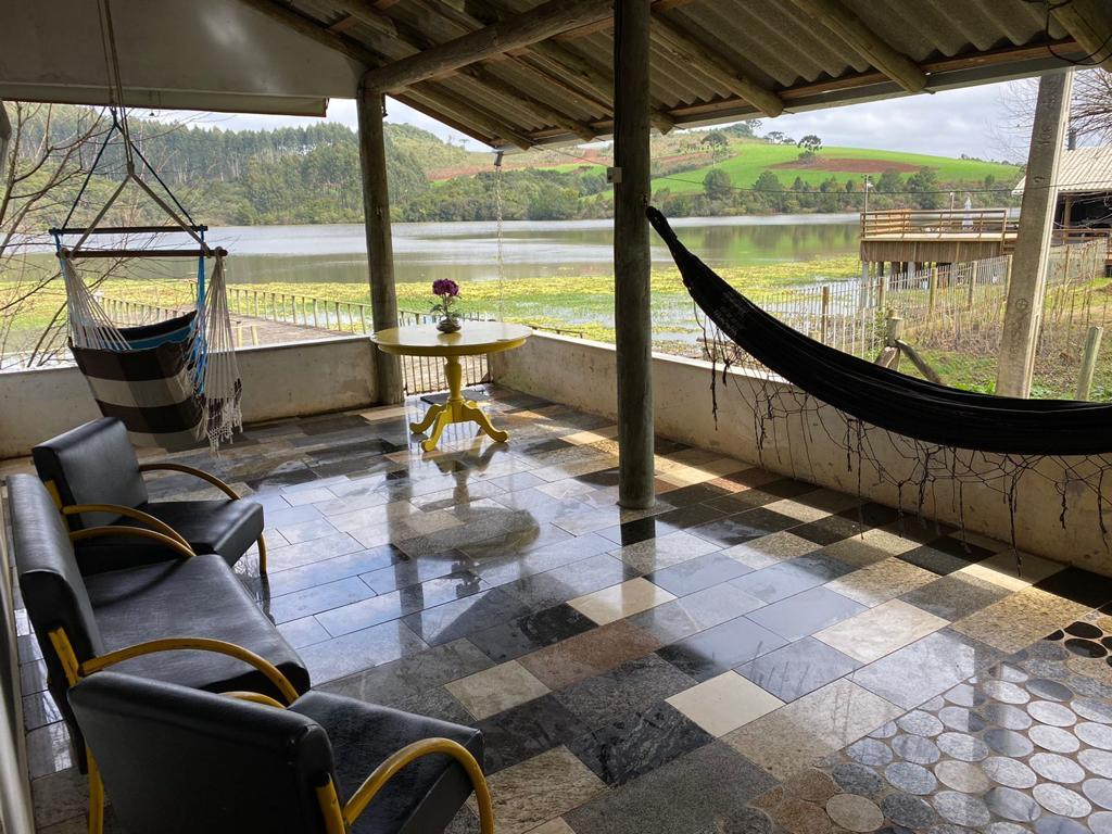 A covered patio features a round table surrounded by simple chairs, complemented by two hammocks. The polished stone floor adds a touch of elegance, while views of the river and lush greenery create a serene backdrop. Decorative elements include soft lighting along the walls.