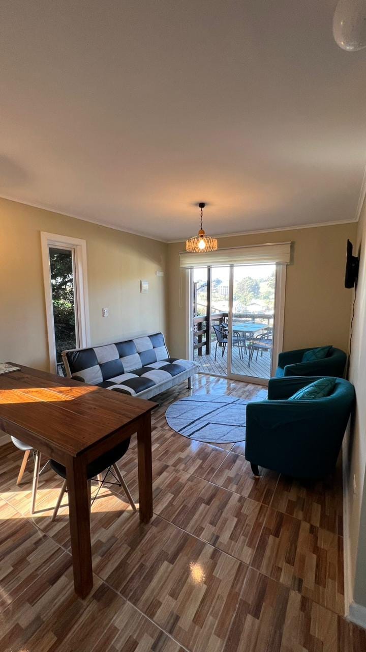 A cozy living area is highlighted with a comfortable seating arrangement that includes a black and white sofa and two teal chairs. A wooden dining table is positioned nearby, with a bright area rug beneath. Natural light enters through sliding glass doors, enhancing the space.