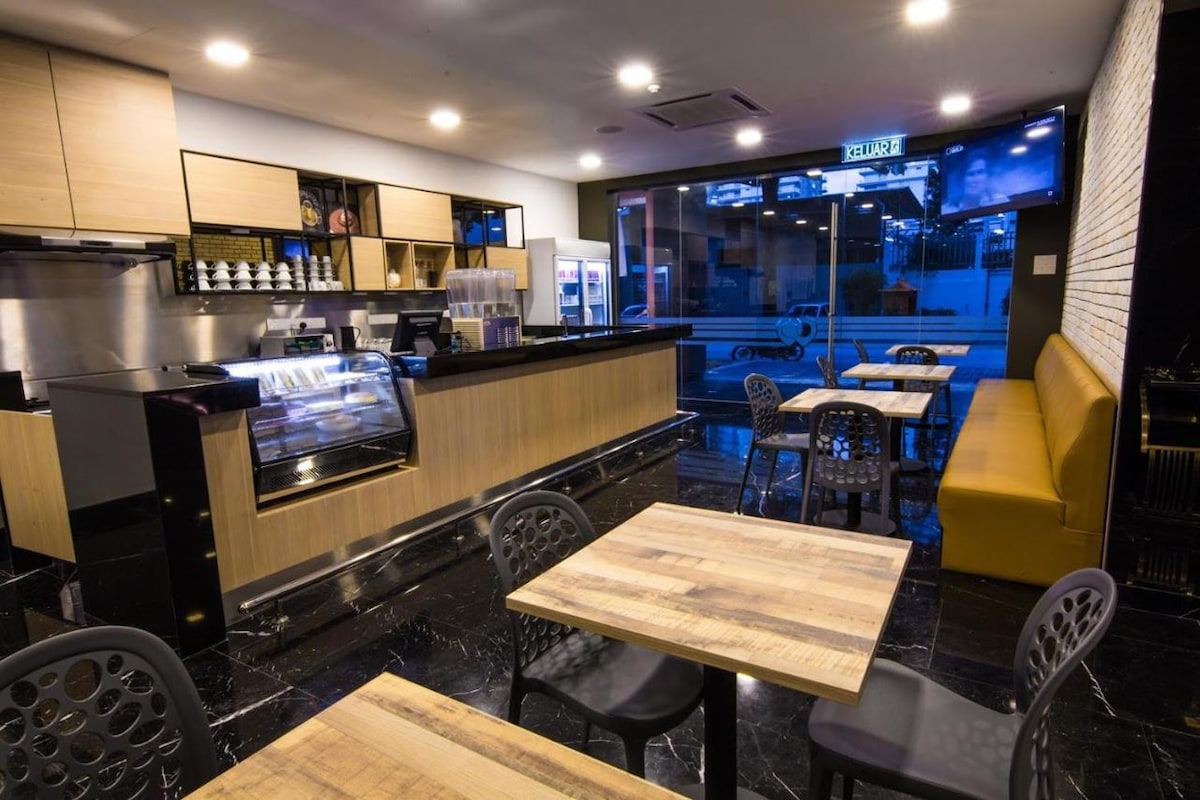 A modern café space is characterized by sleek wooden counters and comfortable seating. The area features several wooden tables surrounded by black chairs, with a spacious counter showcasing various beverages. Large windows allow natural light to fill the room, creating an inviting environment.