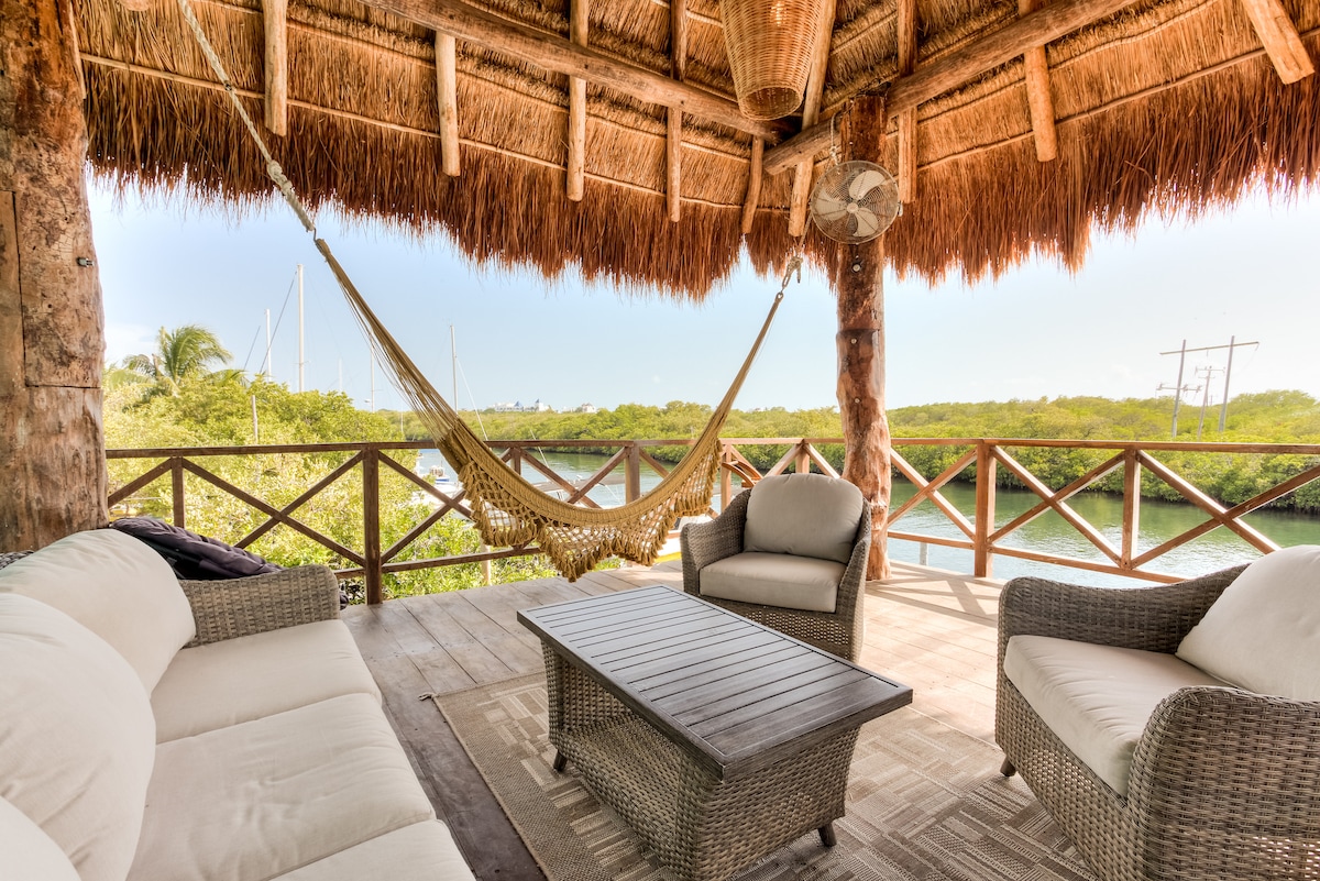 A shaded outdoor seating area features a comfortable sofa and two chairs made of wicker, arranged around a wooden table. A hammock hangs gracefully from the structure's rafters, surrounded by lush greenery and water views, providing a serene space for relaxation.