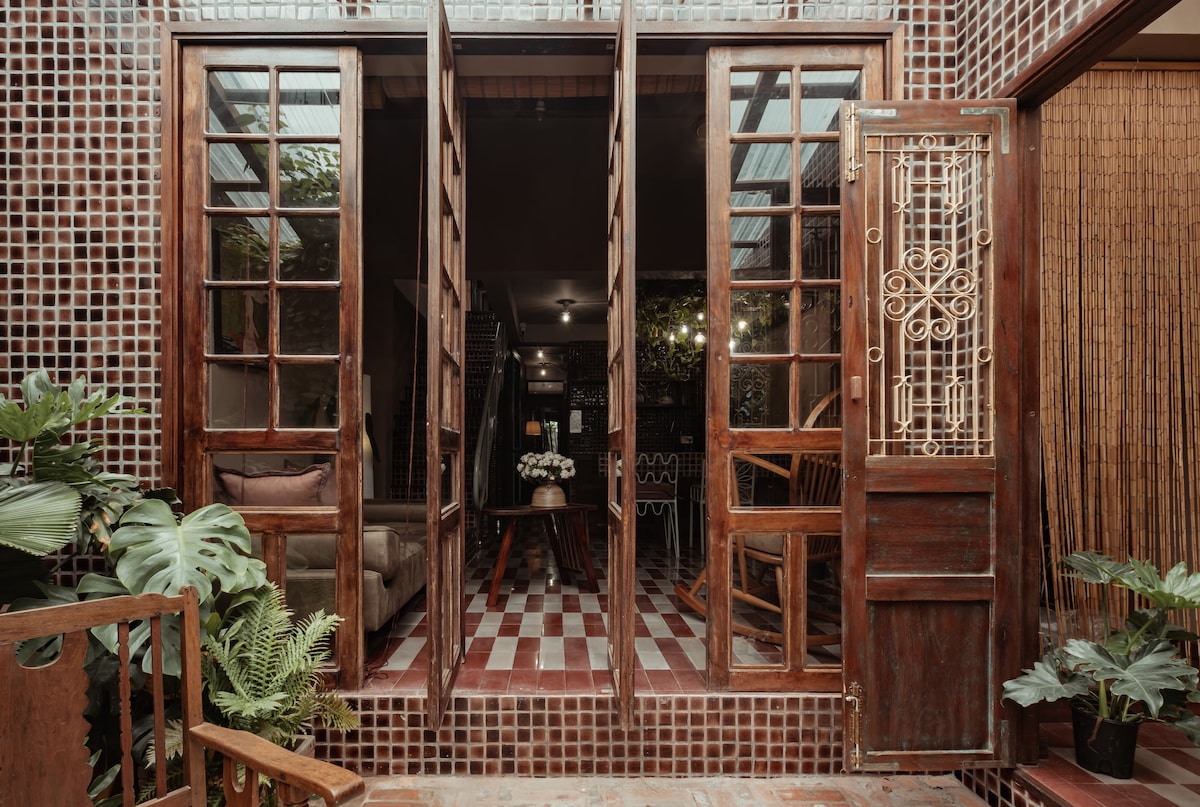 A charming entrance showcases wooden double doors opening to a vibrant interior. The tiled floor features a mix of red and white patterns. Lush green plants are positioned on either side, enhancing the inviting atmosphere of the space.