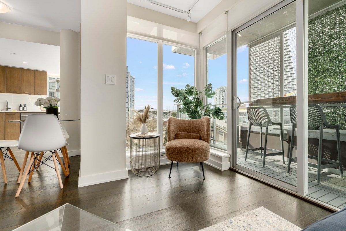 A bright living space features a cozy armchair paired with a small side table. Large glass doors invite natural light and offer views of the balcony, adorned with plants. The adjacent dining area showcases a table with modern chairs, enhancing the open-concept layout.
