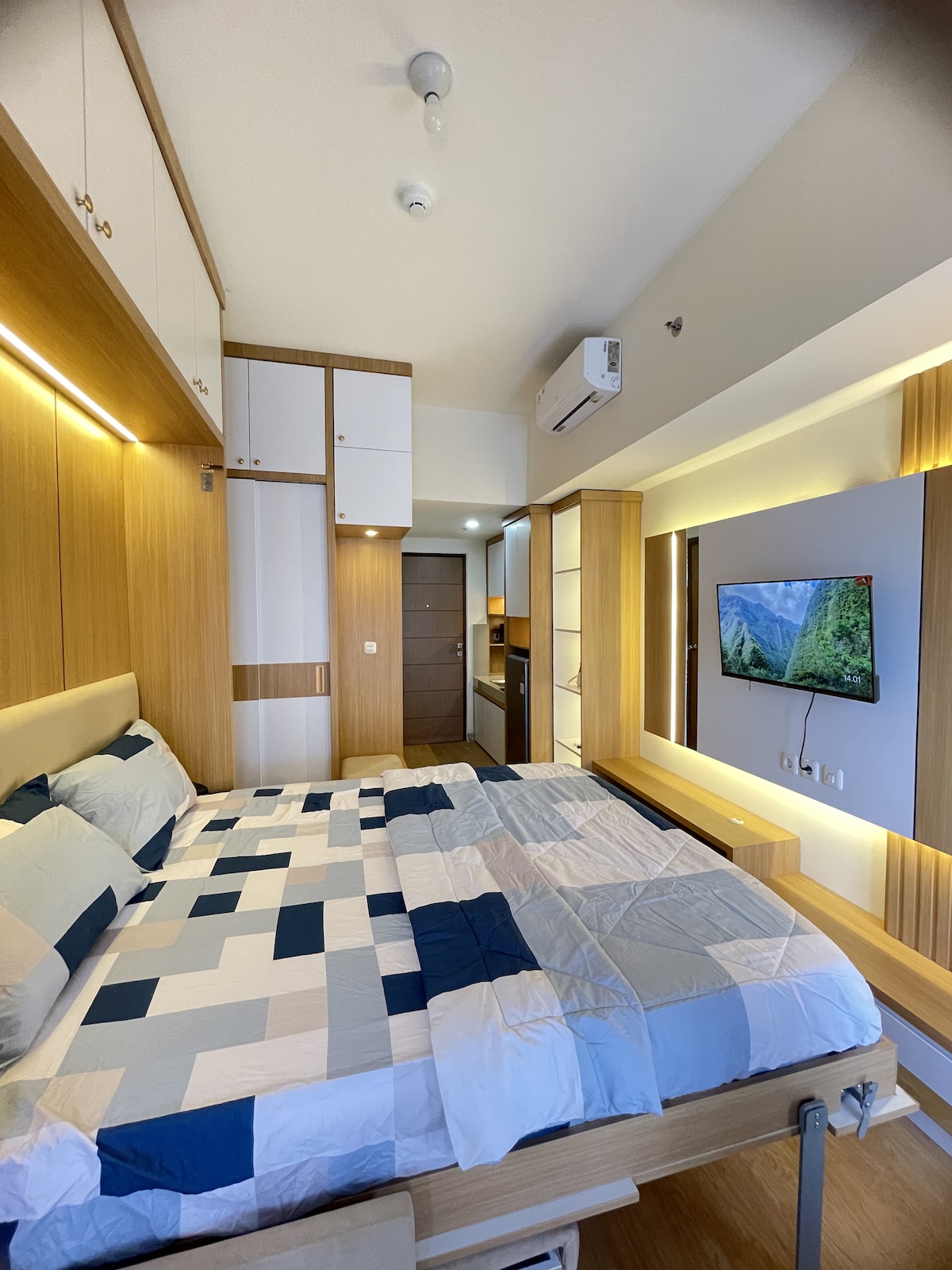 A spacious room features warm wood accents and a queen bed with a geometric patterned blanket. A flat-screen TV is mounted on the wall opposite, and built-in cabinets provide ample storage. Light from the window enhances the natural tones of the décor.