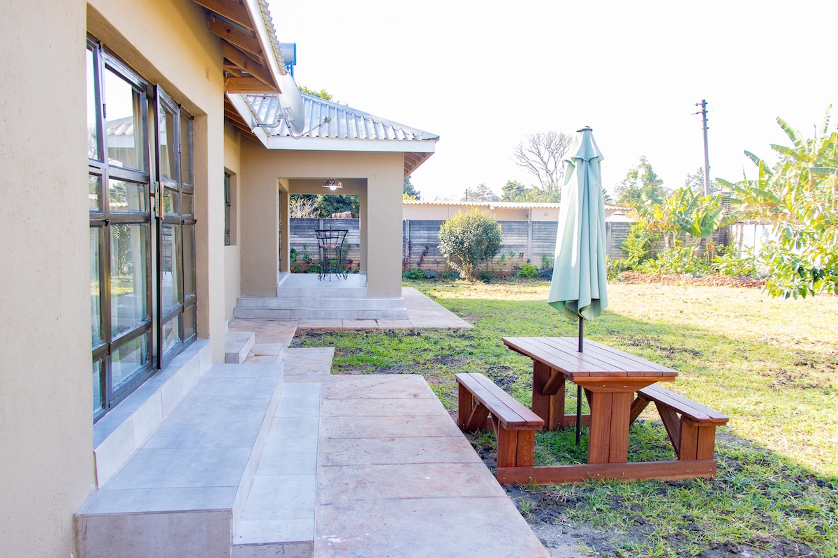 An outdoor space features a wooden picnic table with attached benches and a large umbrella, surrounded by a grassy area. A nearby patio leads to the entrance of the house, accompanied by large windows that provide natural light to the interior.