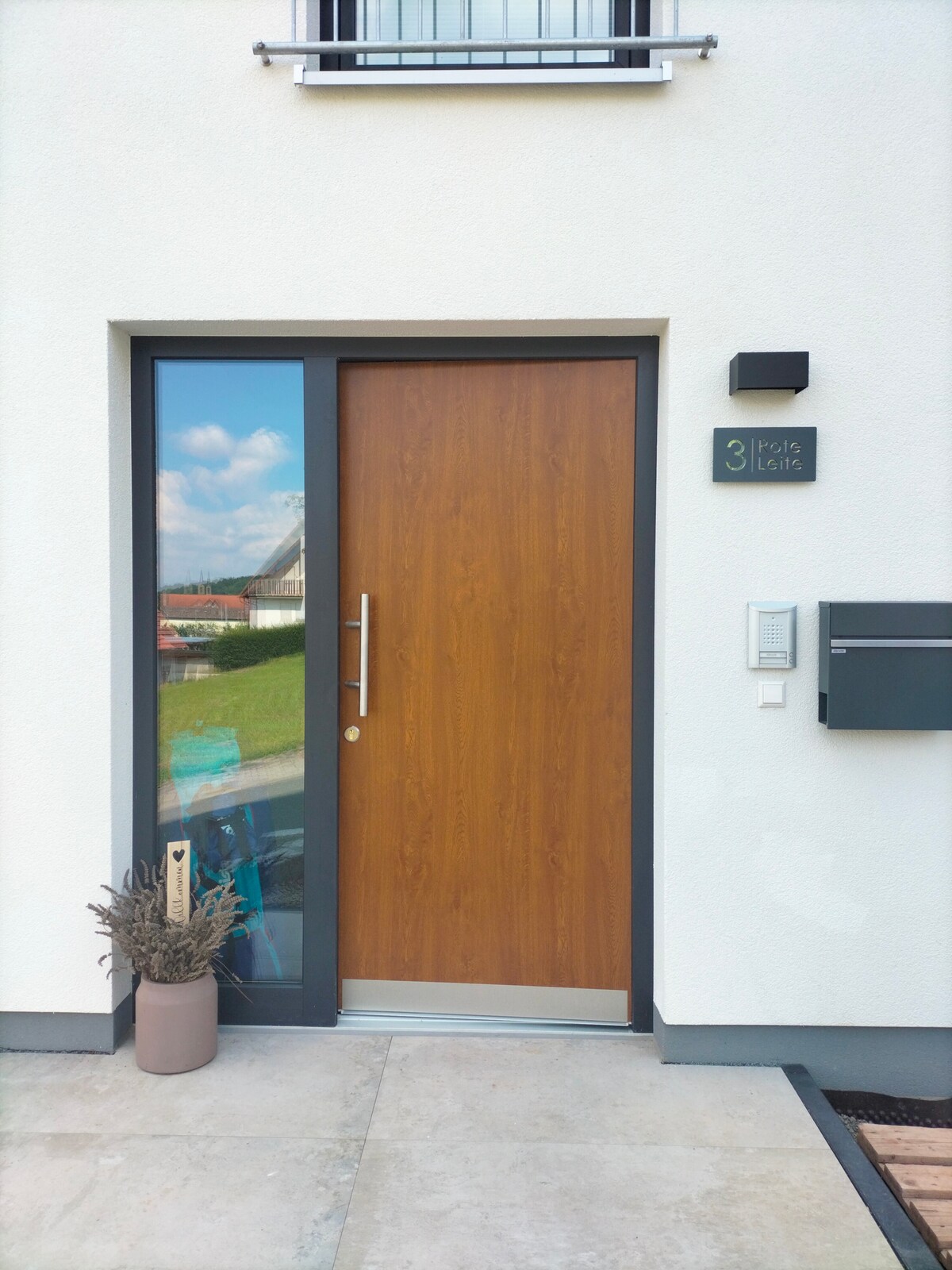 The entrance features a modern wooden door, framed by a sleek grey border. A large window beside the door reflects the surrounding greenery and sky. A small potted plant adorns the entrance, enhancing the clean and minimal aesthetic of the exterior.