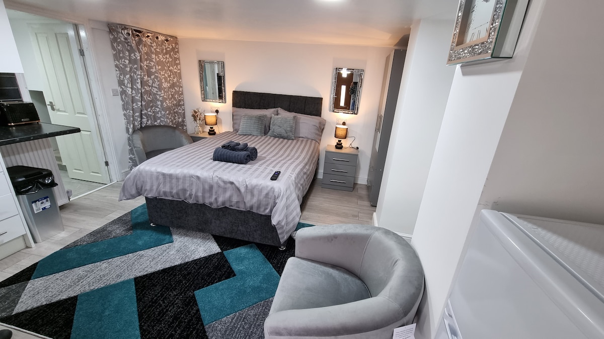 The spacious studio features a comfortable double bed with neutral bedding and two bedside lamps. A cozy armchair sits beside a modern area rug with geometric patterns. Mirrors on the walls reflect light, accentuating the contemporary decor of the room.