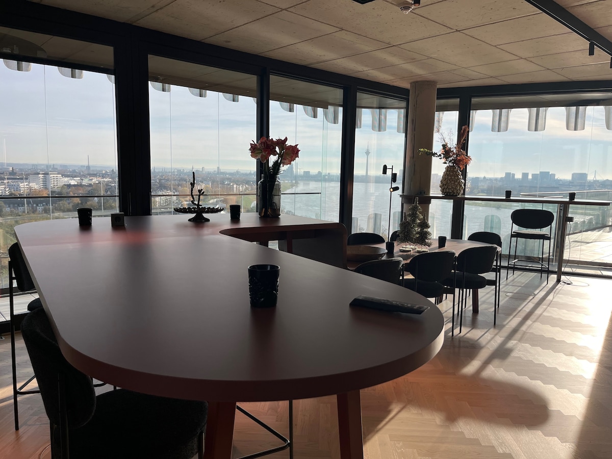 An open dining area features a large, curved table surrounded by black chairs. Natural light floods in from the floor-to-ceiling windows, offering a view of the skyline and water. A vase with flowers adds a touch of color to the space.