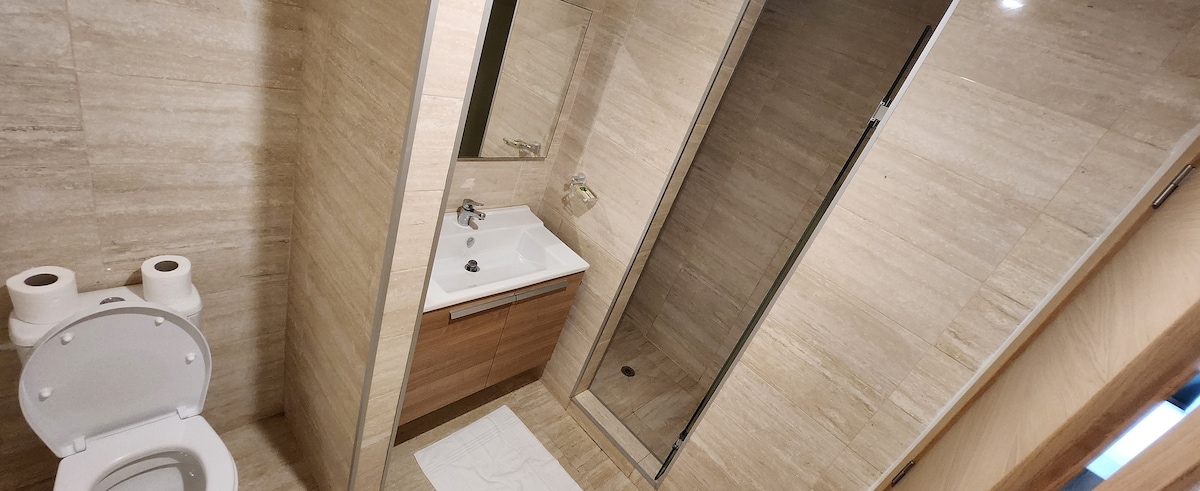 The bathroom features tiled walls and a spacious shower area, separated by glass panels. A modern sink with a wooden vanity is positioned adjacent to the shower, and two rolls of toilet paper are neatly placed beside the toilet for convenience.