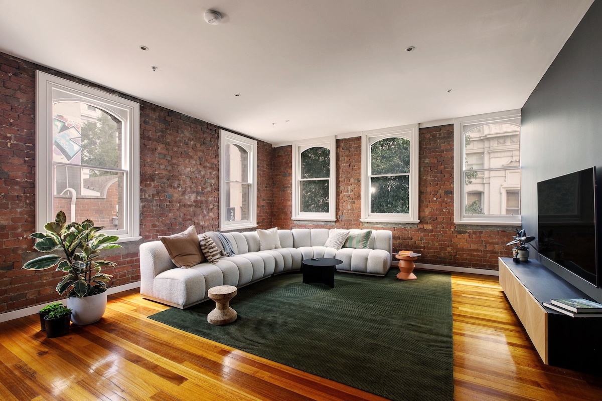 A spacious living area features large windows that allow for natural light to fill the room. Exposed brick walls add texture, while a white sectional sofa is complemented by a green area rug. A small plant and minimalist furniture pieces complete the inviting layout.