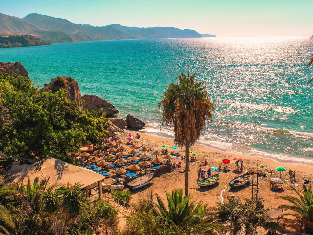 A sandy beach stretches along the shore, bordered by lush greenery and palm trees. Colorful umbrellas provide shade for beachgoers, with small boats anchored nearby. Gentle waves lap against the sand, while distant hills frame the serene coastline under a bright, clear sky.