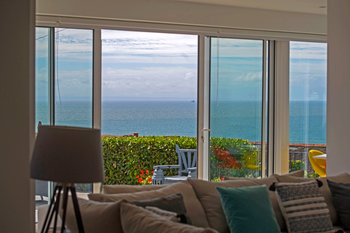 Large glass doors provide a wide view of the sea, framed by greenery. A cozy sofa is positioned within the open-plan living area, allowing for comfortable relaxation while enjoying the panoramic ocean scenery.