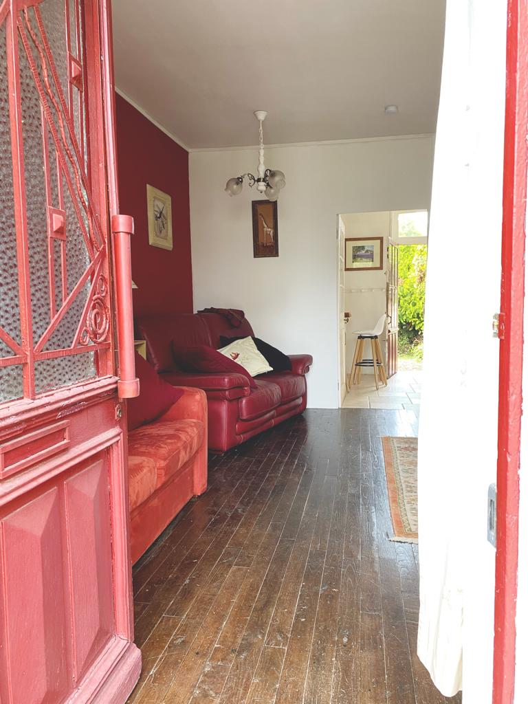 A welcoming entryway leads into the living room, featuring a cozy arrangement of red sofas placed against a red accent wall. Natural light filters in from an open door, hinting at the adjoining garden. A wooden floor extends throughout the space, enhancing its traditional charm.