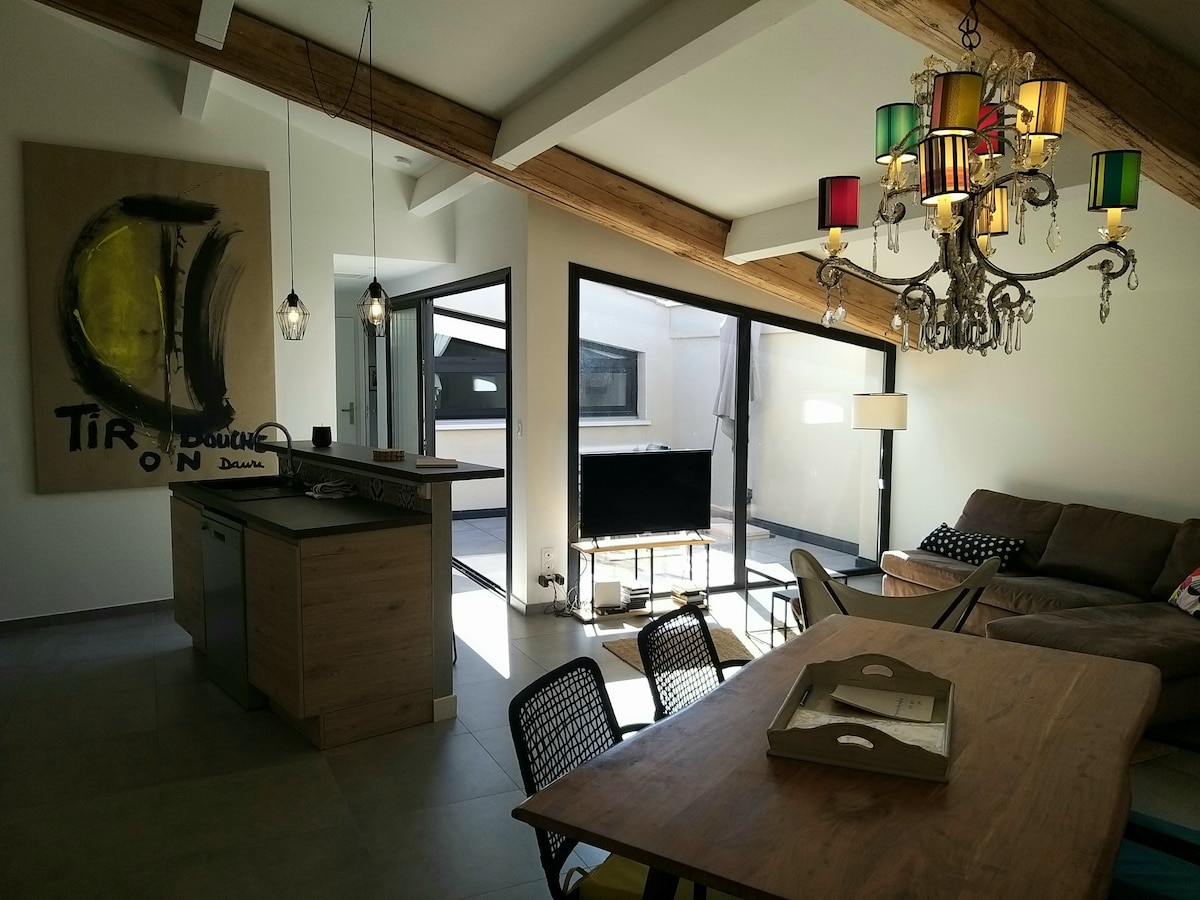 An open living space is highlighted by wooden beams and contemporary furnishings. A kitchen area features sleek cabinetry and modern appliances. Large glass doors create a connection to a private outdoor area, where natural light enhances the inviting atmosphere of the room.