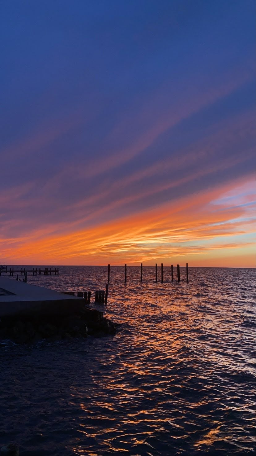 A vibrant sunset casts a warm glow over the water, blending hues of orange, purple, and pink. Silhouetted pilings rise from the bay, while gentle waves reflect the colorful sky. A small dock is visible in the foreground, inviting guests to enjoy the serene view.