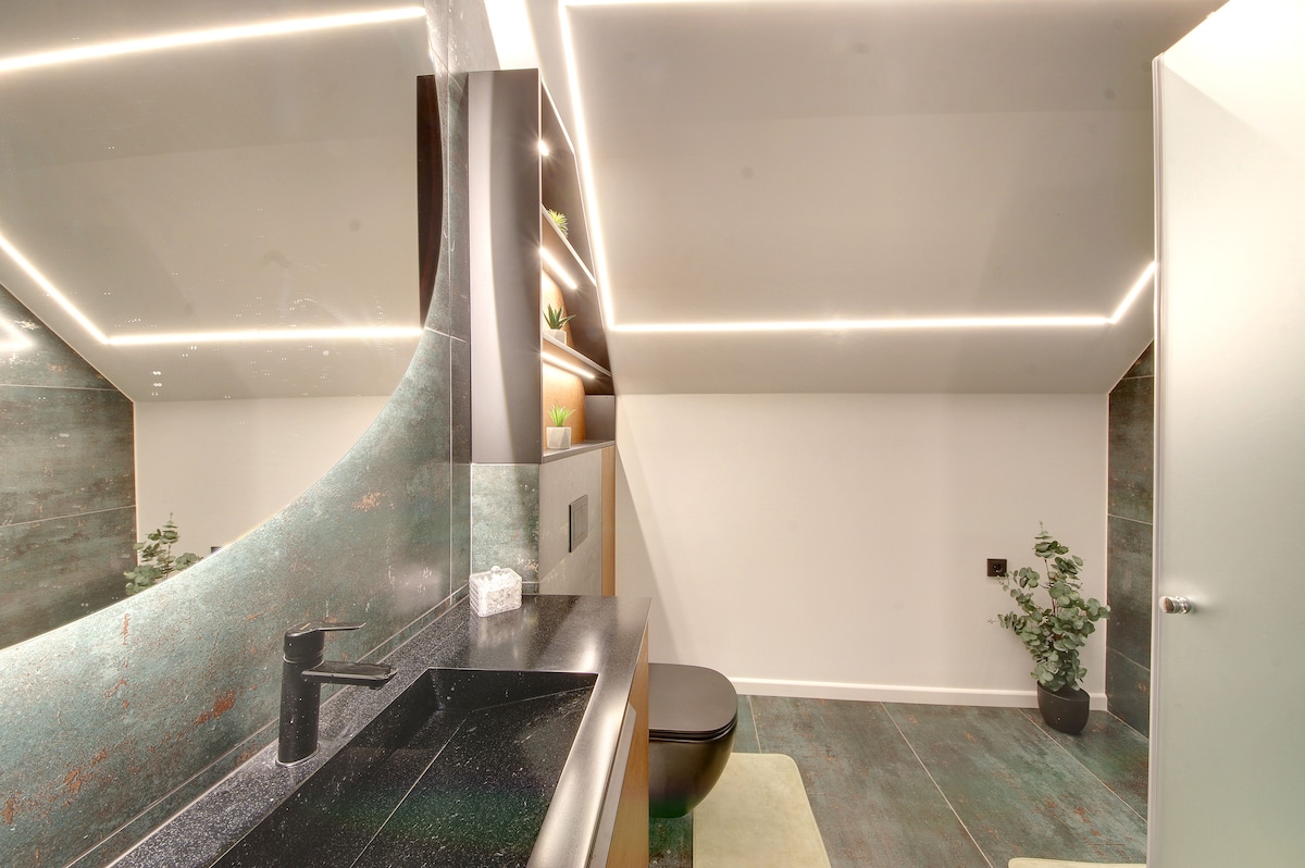 A modern bathroom features sleek, dark cabinetry and a black sink against textured green walls. Soft lighting highlights the space, while a large mirror enhances the feeling of openness. A small potted plant adds a touch of greenery to the contemporary design.