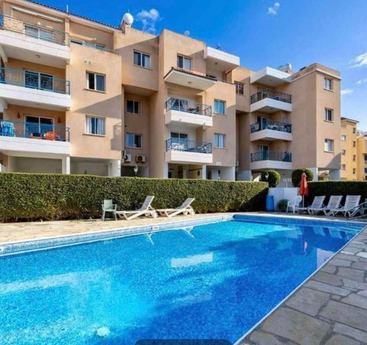 A common swimming pool is surrounded by well-maintained greenery and features a sunbathing area with lounge chairs. The beautifully designed apartment complex rises in the background, showcasing multiple balconies and bright facades under a clear blue sky.