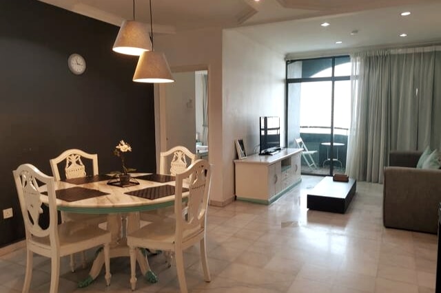 A dining area features a round table surrounded by four elegant chairs, positioned against a dark accent wall. Natural light filters through large windows that lead to a balcony, while a cozy living area is visible, showcasing a modern sofa and television unit.
