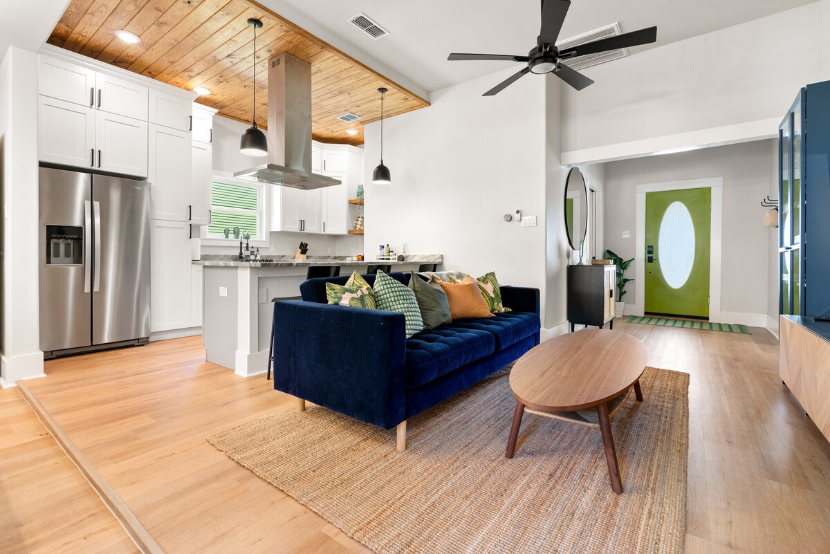 An open-concept living area features a navy blue sofa adorned with colorful cushions, a round wooden coffee table, and a natural fiber rug. The modern kitchen, equipped with stainless steel appliances and white cabinetry, is illuminated by pendant lights, while a green accent door provides a vibrant entryway.