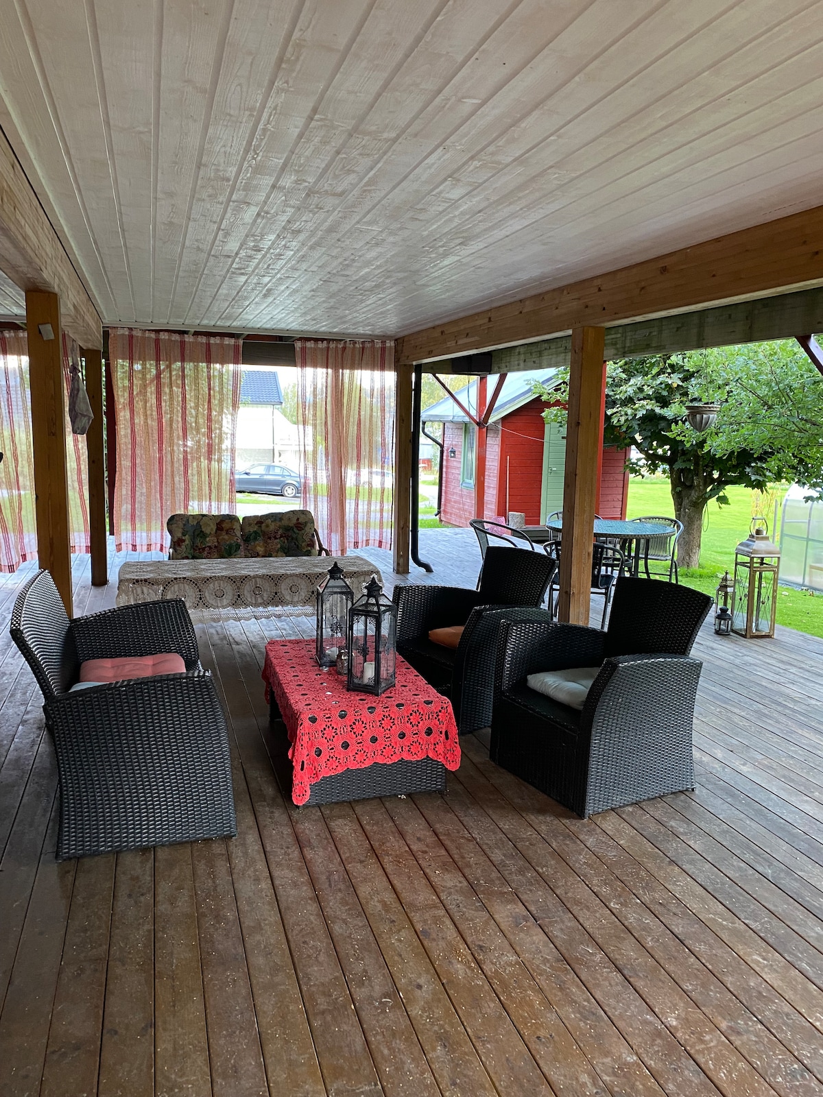 A covered outdoor seating area features a mix of wicker chairs and a patterned sofa, arranged around a red tablecloth-covered table. Lanterns add warmth to the space, while sheer curtains provide a sense of privacy, and greenery from the surrounding yard is visible.