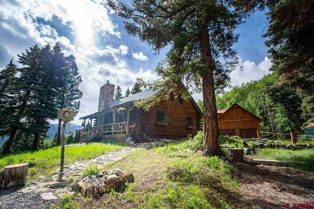 The rustic log cabin features a stone chimney and a welcoming front porch. An inviting pathway made of stones leads through green grass to the entrance. Surrounding pine and aspen trees enhance the natural setting, while distant mountain views are visible under a partly cloudy sky.