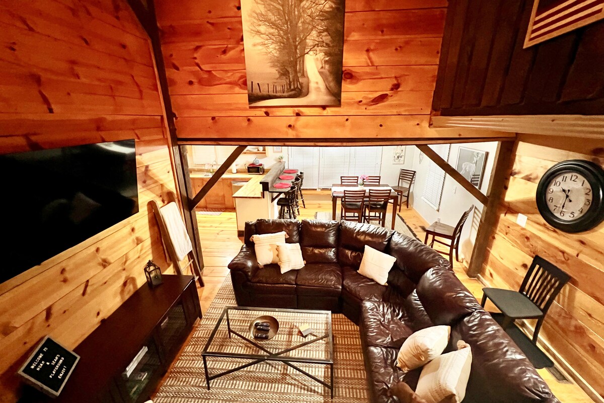 An expansive living area features a large leather sectional sofa arranged around a glass coffee table. Rustic wooden walls add warmth, while a large framed artwork of a tree is mounted above. A dining space and kitchen are visible in the background.